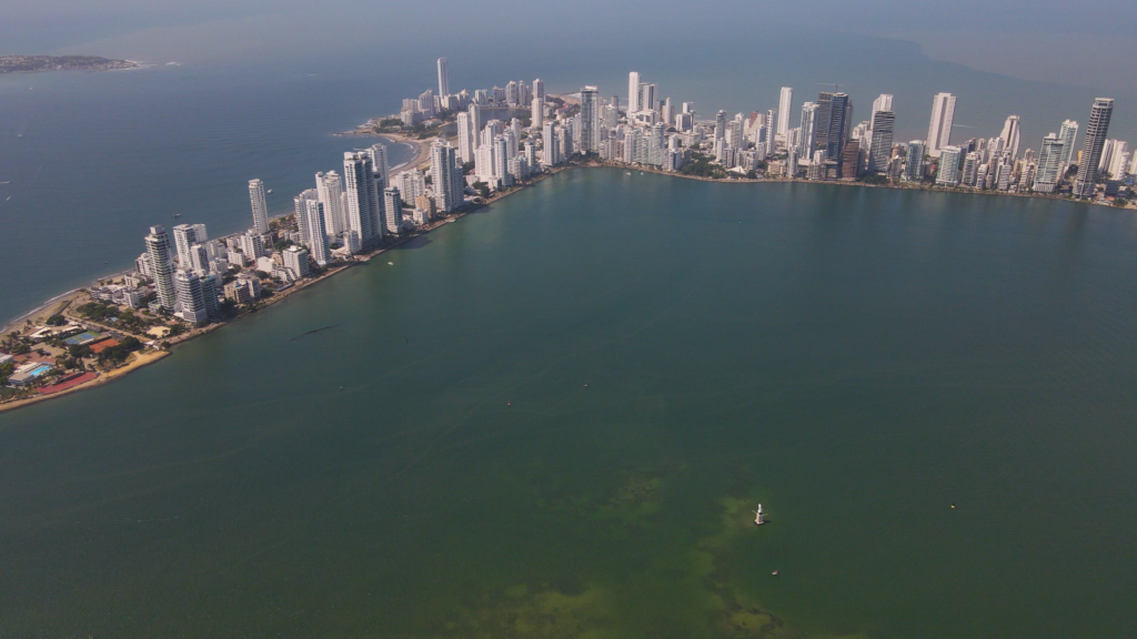 Aerial view of a modern coastal cityscape with numerous high-rise buildings surrounded by an ocean bay and a greenish sea.