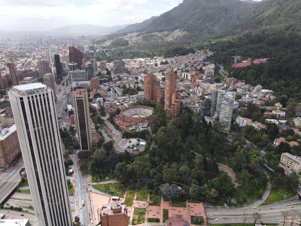 Vista aérea de un paisaje urbano con edificios altos, una estructura circular parecida a una plaza de toros y una zona boscosa junto a una montaña. Por toda la ciudad se ven carreteras y edificios más pequeños.