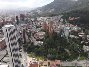 Aerial view of a cityscape with tall buildings, a circular structure resembling a bullring, and a forested area by a mountain. Roads and smaller buildings are visible throughout the city.