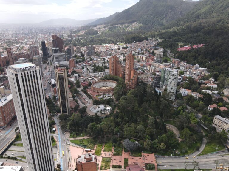 Aerial view of a cityscape with tall buildings, a circular structure resembling a bullring, and a forested area by a mountain. Roads and smaller buildings are visible throughout the city.