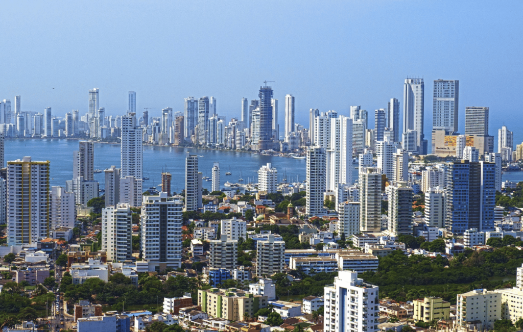 Aerial view of Cartagena's coastal cityscape, with numerous skyscrapers and residential buildings, framed by the historic Walled City and the shimmering waters stretching towards the Rosario Islands in the background.