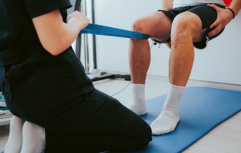 A person assists another in performing knee exercises with a resistance band on a mat, focusing on rehabilitation after a knee replacement, highlighting the possibilities of medical tourism in Colombia.