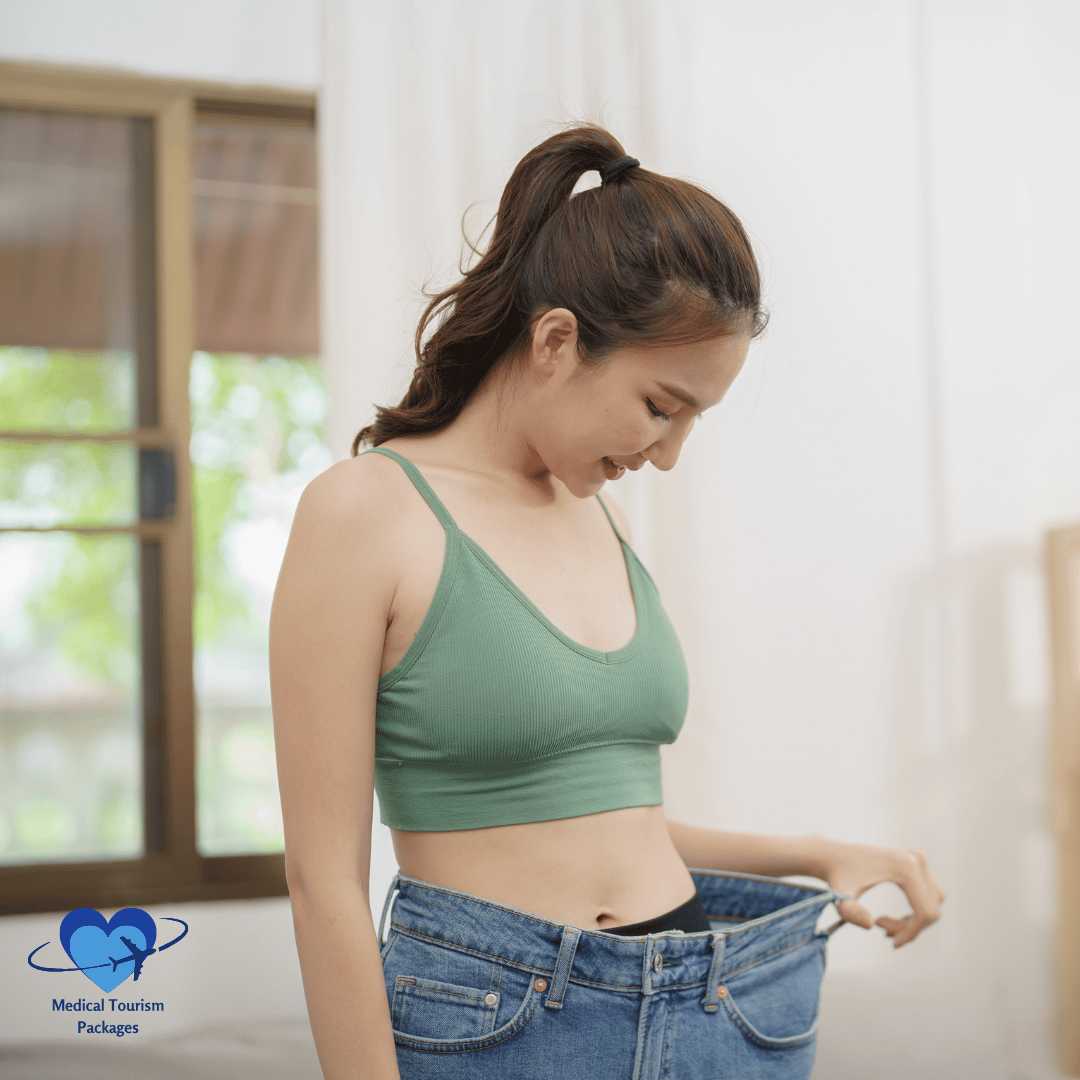 A person in a green tank top looks down at their loose-fitting jeans, holding the waistband—a testament to successful weight loss. A window with blinds is in the background, letting in soft, natural light.