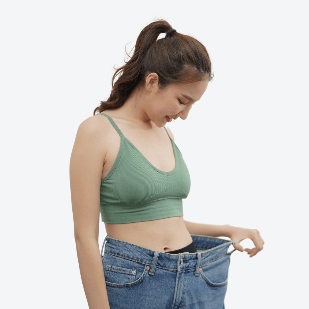 A woman in a green tank top holds out the waistband of oversized jeans, smiling and looking down.