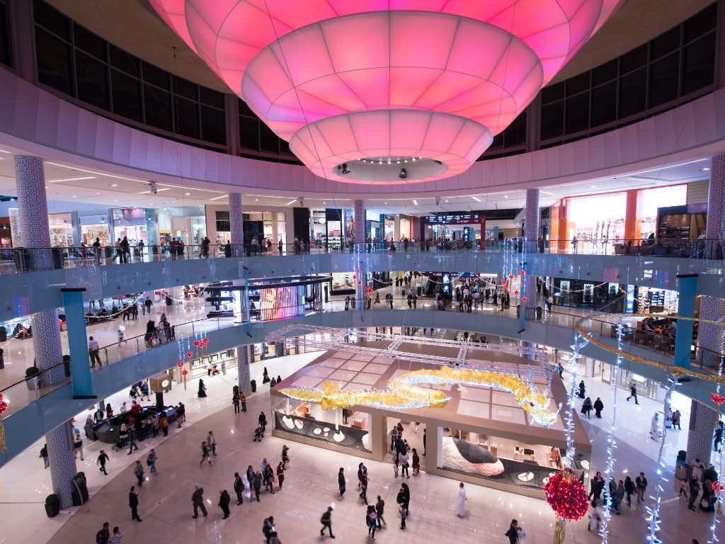 Spacious shopping mall interior with a large, circular pink ceiling light invites you to unwind. People walk on multiple levels, exploring shops and modern decor throughout. A sense of rejuvenation fills the air, transforming your visit into a Caribbean Renewal experience.