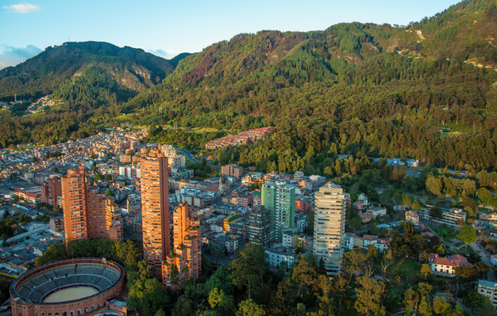 Aerial view of a city with tall buildings, a circular stadium, and surrounding forested hills under a blue sky, with the historic charm of La Candelaria nestled below and Monserrate majestically overlooking the bustling expanse.