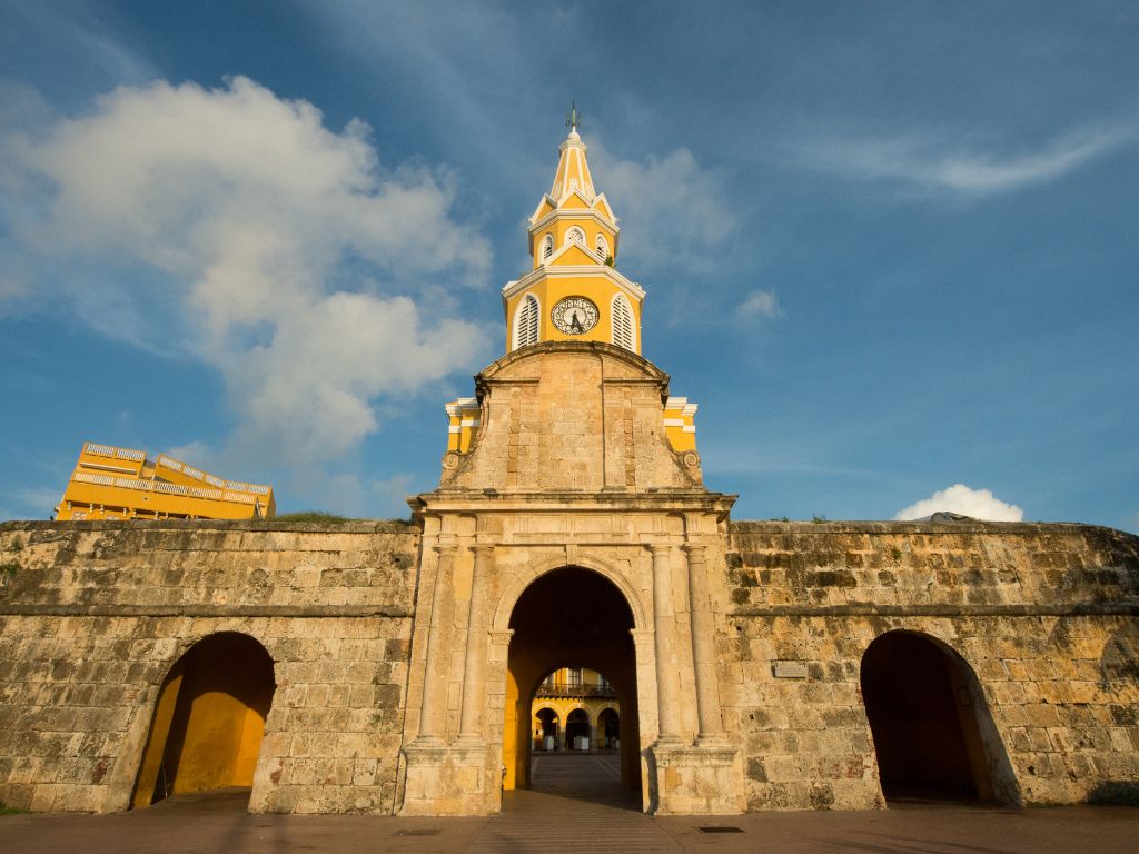 La torre del reloj amarilla con chapitel corona la rejuvenecida e histórica estructura de piedra de la puerta, enmarcada en un cielo azul caribeño salpicado de nubes dispersas.