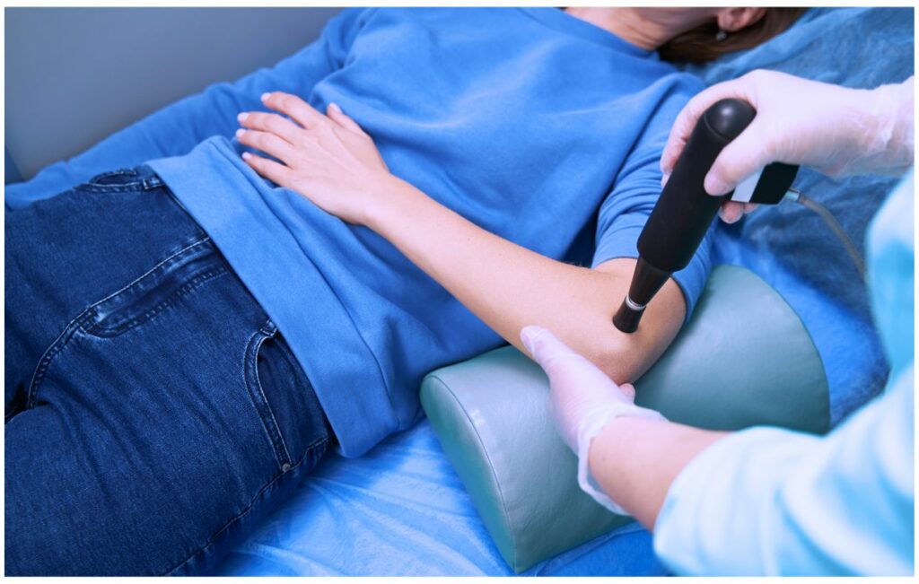 Primer plano de un profesional médico con guantes blancos tratando el brazo de un paciente con un dispositivo terapéutico manual. El paciente lleva ropa azul y está tumbado en una cama médica gris mientras recibe un tratamiento focalizado en un entorno clínico.