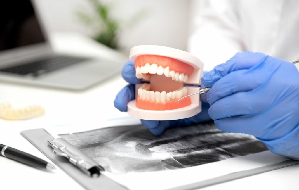 Un dentista con guantes azules utiliza una herramienta dental para examinar un modelo de diente sobre una radiografía y un portapapeles en el escritorio, asegurando el camino hacia una sonrisa perfecta.