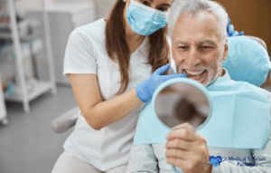 Un hombre mayor en un sillón dental sostiene un espejo de mano, con una amplia sonrisa tras ponerse implantes dentales, mientras un profesional de la odontología con mascarilla y guantes está a su lado.