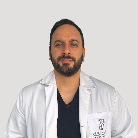 Dr Tirupati a beard wearing a white lab coat over a dark shirt stands against a light gray background.