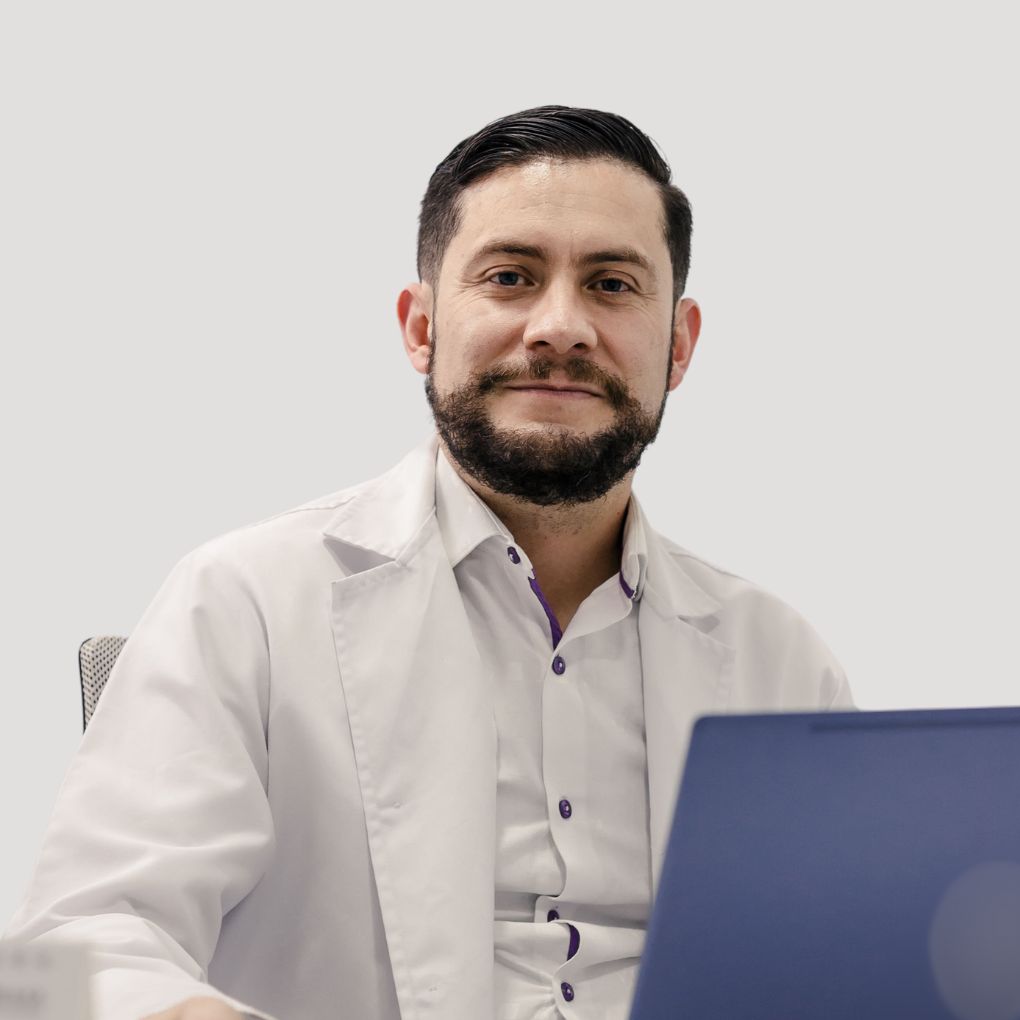 A person with a beard, wearing a white coat, sits in front of a blue laptop, looking at the camera with a light background.