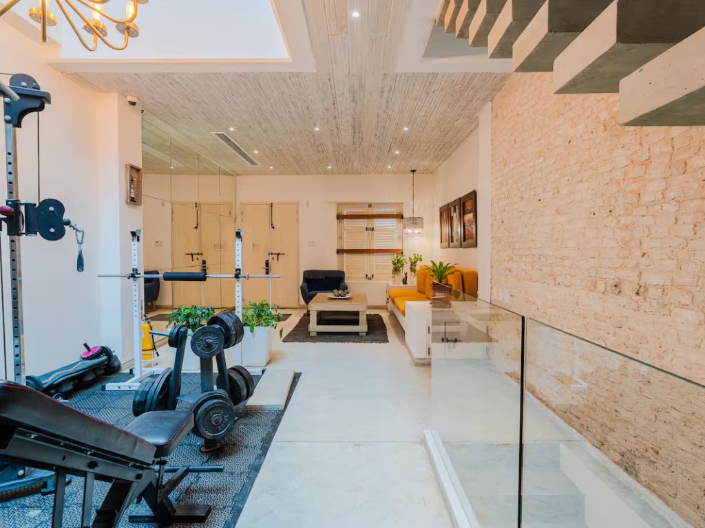 Unwind in a home gym and living area featuring exercise equipment, a white sofa, and vibrant yellow seating. An exposed brick wall and ceiling lights enhance the modern interior, creating a perfect space for rejuvenation.