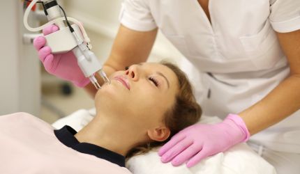 A person enjoys a rejuvenating facial treatment with a handheld device, wearing a pink bib, as the practitioner, donning gloves, gently supports their head.