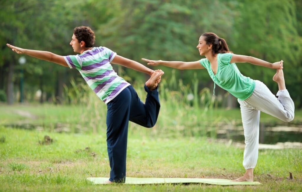 Dos personas practican yoga al aire libre, demostrando posturas de equilibrio con una sola pierna en un entorno natural con árboles y agua, representando actividades de rehabilitación postoperatoria en el entorno curativo de Colombia.