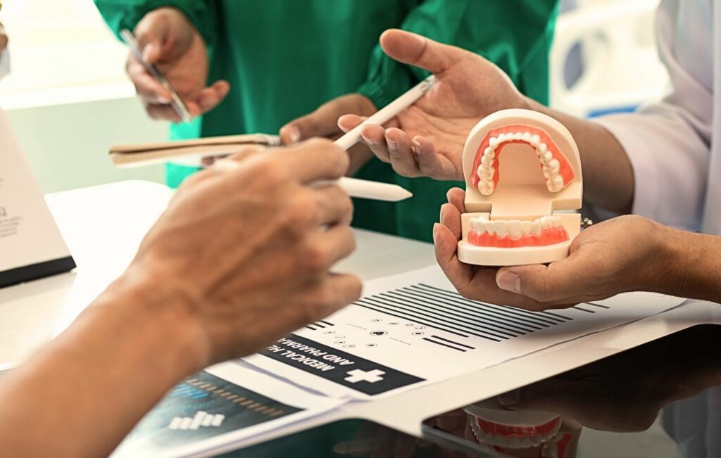 Profesionales médicos con atuendo verde y blanco discutiendo planes de tratamiento mientras sostienen modelos dentales y documentos, con las manos señalando papeles y mostrando un modelo de dentadura completa durante una reunión de consulta.