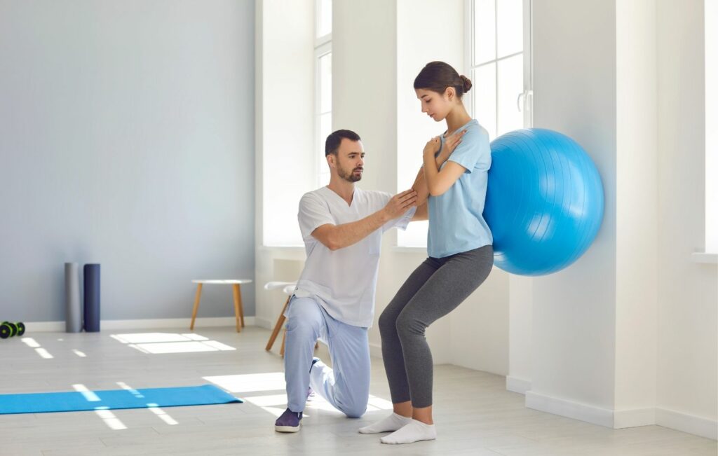 Fisioterapeuta con atuendo blanco arrodillado junto a una paciente con camisa azul claro y leggings grises, guiándola a través de un ejercicio con pelota de estabilidad contra una pared blanca en una sala de rehabilitación iluminada por el sol con equipos de ejercicio visibles en el fondo.