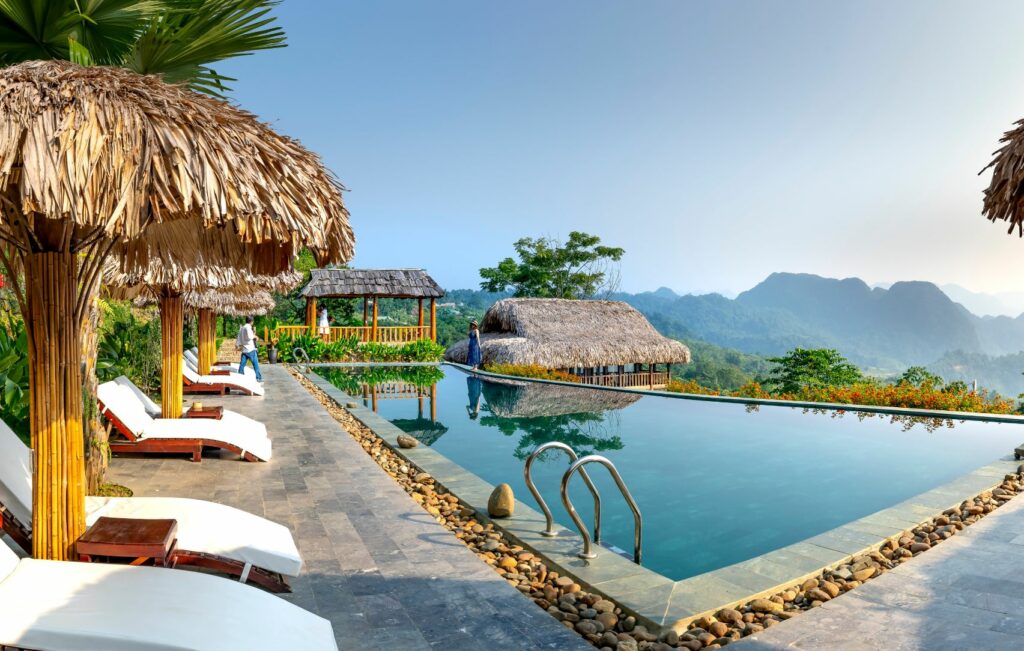 La piscina infinita con borde de piedra y tumbonas, rodeada de cabañas de paja, ofrece una muestra de lujo asequible. Tiene vistas a las verdes colinas y montañas de Panamá bajo un cielo azul despejado.