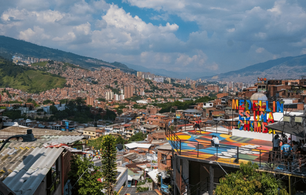 A vibrant cityscape of Medellín, Colombia, showcases its mountainous terrain and a colorful rooftop sign reading "Medellin es una chimba." In the heart of Comuna 13, the energy is palpable, much like during the exhilarating Flower Festival.