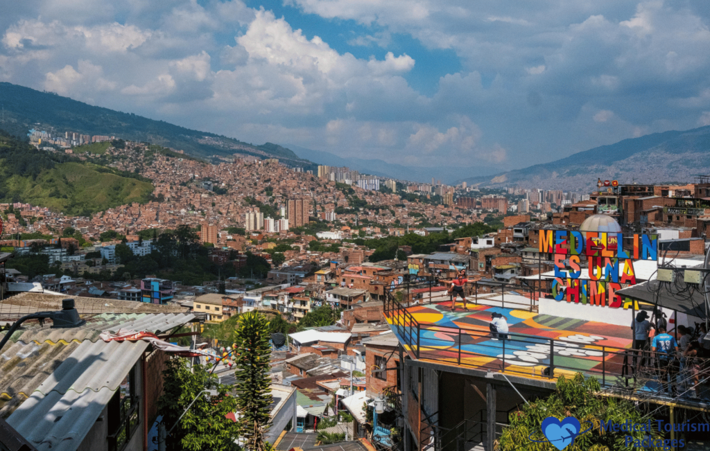 A vibrant urban neighborhood on a hillside in Colombia, featuring a colorful sign reading "Medellín Es Una Chimba," is set against a backdrop of mountains and a partly cloudy sky. This picturesque spot is among the top tourist attractions for those exploring with their travel guide.