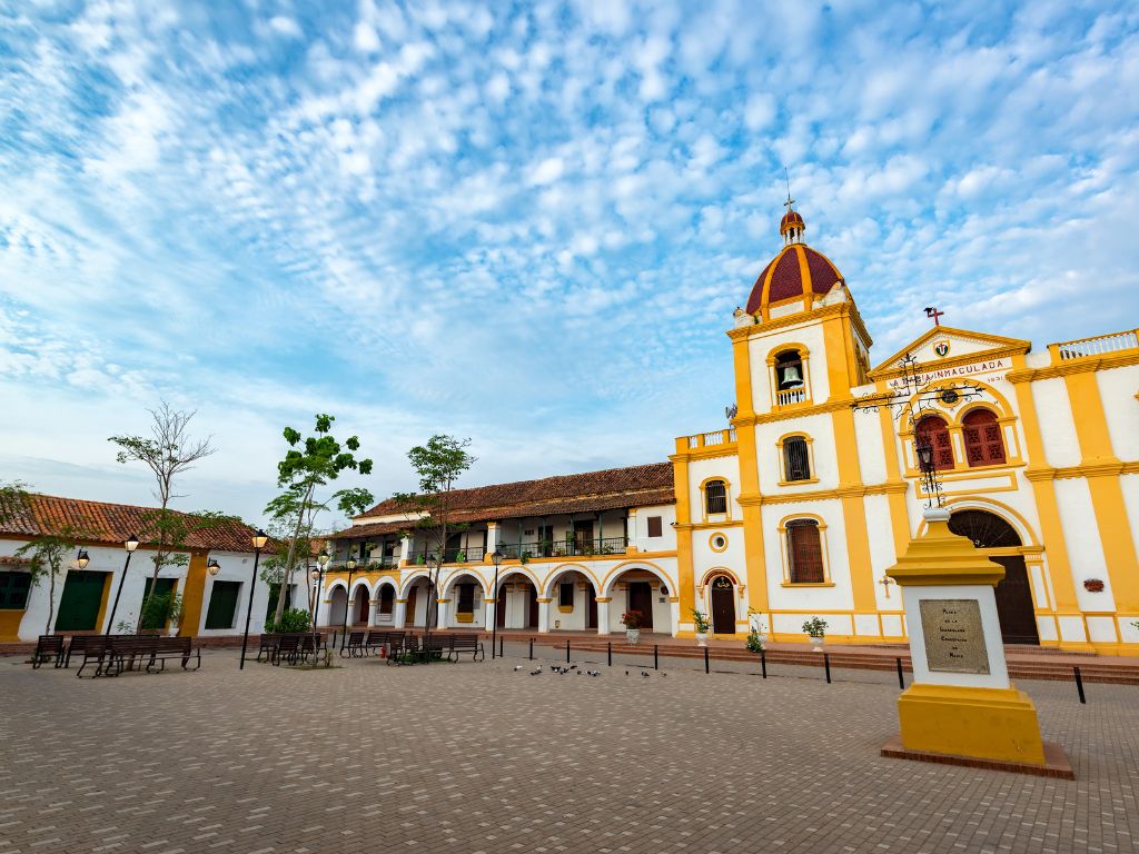 Plaza de estilo colonial con una iglesia amarilla y blanca, un edificio arqueado y un monumento en primer plano bajo un rejuvenecedor cielo azul nublado, que evoca el vibrante espíritu del Caribe.