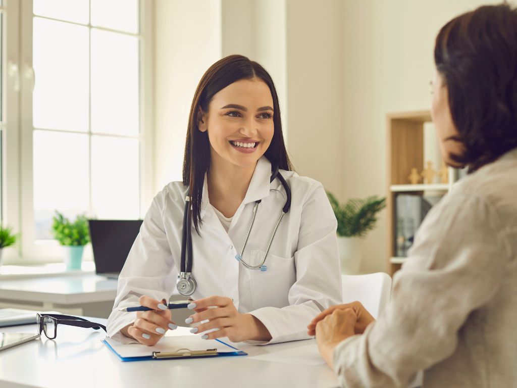 A doctor in a white coat and stethoscope smiles, bringing a sense of rejuvenation as they talk to a patient across a desk in a bright office.