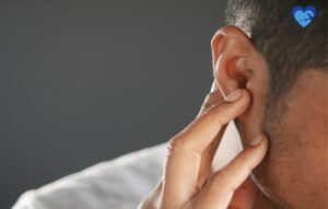 Primer plano de una persona con el pelo corto presionando suavemente su oreja, posiblemente pensando en levantarse la oreja, con un fondo borroso y un icono de un corazón azul en la esquina superior derecha.