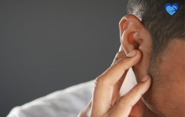 Close-up of a person with short hair gently pressing their ear, possibly considering ear lifts, with a blurred background and a blue heart icon in the top right corner.