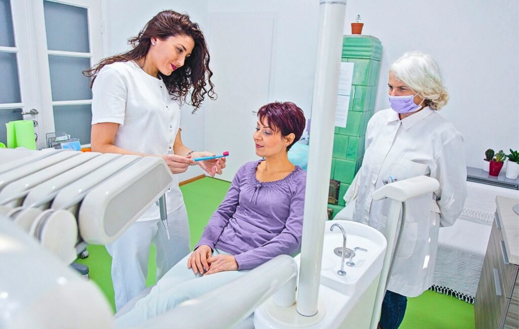 A female dentist demonstrates a toothbrush to a woman in the dental chair, aiming for that perfect smile, as another masked dentist observes. The room, with its green floor and advanced dental equipment, embodies the dedication to smile correction you’d expect in Panama.