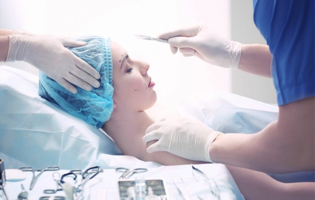 A patient lies on a surgical table, wearing a blue cap, undergoing rhinoplasty surgery. The surgeon in blue scrubs skillfully wields a scalpel, assisted by another person. Surrounded by precision tools, they aim for natural results and affordable precision in every incision.