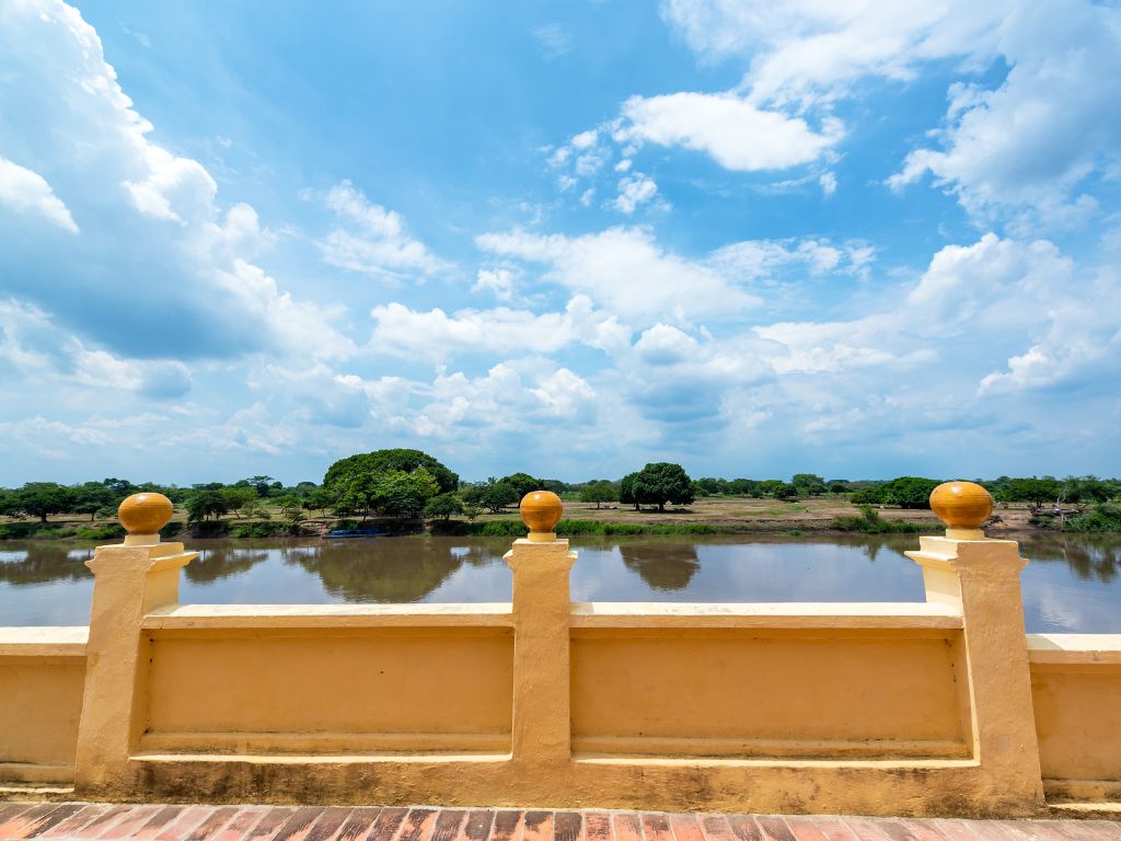 Yellow stone railing overlooking a calm river invites you to unwind as trees grace the opposite bank under a cloudy sky.