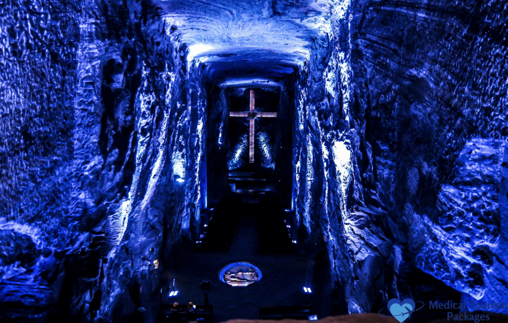 A dimly lit underground cavern in Colombia, with ethereal blue lighting, features a large illuminated cross at the far end, serving as a captivating guide through one of the nation's unique tourist attractions.