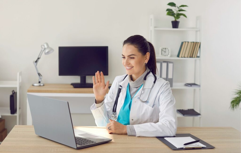 A doctor in a white coat and stethoscope waves at a laptop screen in a medical office lined with shelves, books, and a plant. The virtual consultation is focused on discussing abdominoplasty in Panama, providing patients with expert advice from afar.