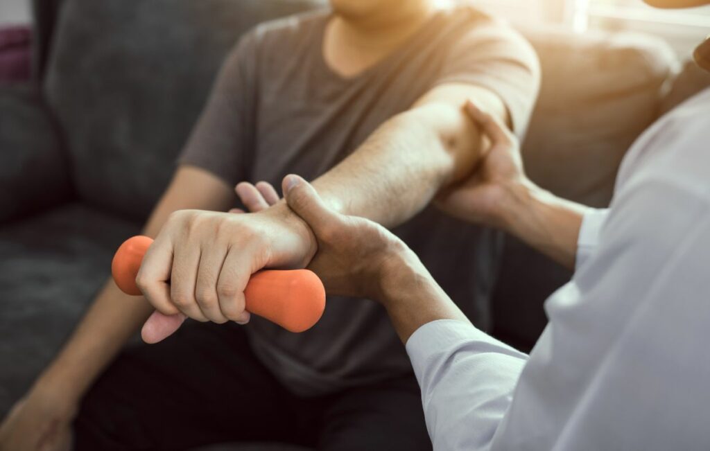 Un terapeuta de medicina deportiva con bata blanca guía a un paciente a través de ejercicios de rehabilitación del hombro, apoyando el movimiento de su brazo mientras el paciente agarra una mancuerna naranja en un entorno clínico bien iluminado con luz natural.