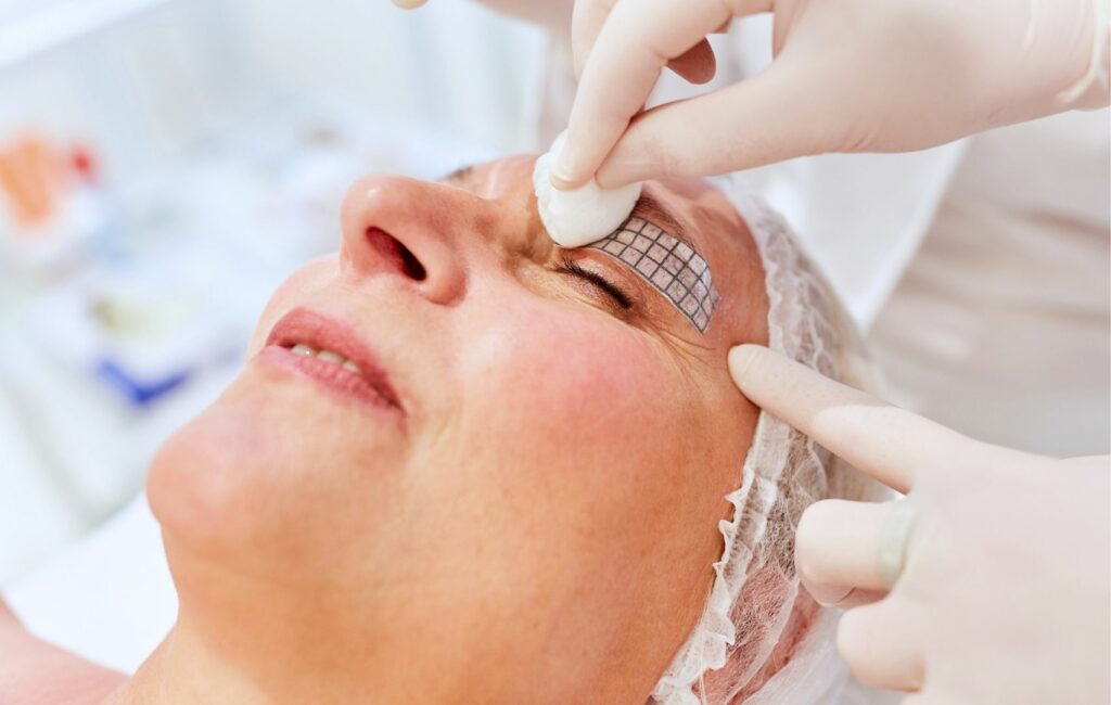 A person in Panama receives a cosmetic eyebrow treatment, donning a hairnet, as a professional applies a grid pattern on the brow area using gloves and a cotton pad.